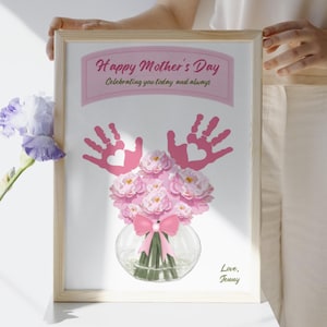 Pode incluir: Uma impressão emoldurada com o tema do Dia das Mães. A obra de arte apresenta impressões de mãos cor-de-rosa, um buquê de flores cor-de-rosa em um vaso de vidro e o texto "Happy Mother's Day". A impressão é segurada por uma pessoa.