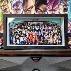 Op de afbeelding: Een ingelijst 3D-kunstwerk met tal van Dragon Ball Z-personages. Het kunstwerk is geplaatst tegen een achtergrond van verschillende karakterportretten. De lijst is zwart en het kunstwerk wordt op een houten oppervlak gepresenteerd.