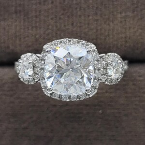 May include: A three-stone engagement ring with a large, square-cut center stone surrounded by a halo of smaller diamonds. Two smaller round stones flank the center stone. The ring is set in a silver band.