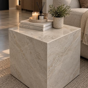 May include: A cube-shaped side table featuring a polished beige and cream marble-look finish with subtle veining. The top surface holds two stacked books titled The Kinfolk Home, a round speckled tray with a candle, a reed diffuser, and a plant.