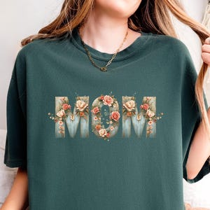 Puede incluir: Sudadera de color verde azulado con la palabra "MOM" en un diseño floral. Las letras son de color azul claro con una textura de madera, adornadas con rosas rosas y crema y vegetación. Se ve un collar dorado.