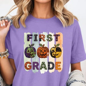 Puede incluir: Una camiseta de color morado claro con un estampado gráfico que incluye el texto FIRST GRADE. El diseño presenta tres calabazas de Halloween en verde, naranja y negro sobre franjas verticales de colores pastel con motivos festivos.