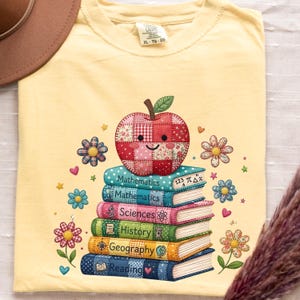 以下が含まれることがあります： 淡い黄色のTシャツで、笑顔のパッチワーク風リンゴが乗った6冊の本の積み重ねが描かれています。本にはMathematics、Sciences、History、Geography、Readingと書かれており、小さな花、ハート、星の模様が添えられています。