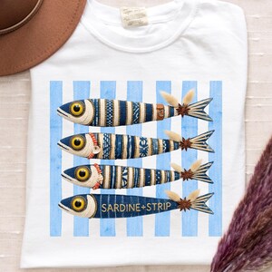 Puede incluir: Una camiseta blanca con un gráfico de cuatro peces estilizados dispuestos horizontalmente sobre un fondo de rayas verticales azul claro. Los peces están decorados con patrones azules y blancos, ojos amarillos y detalles de anís estrellado en las colas.