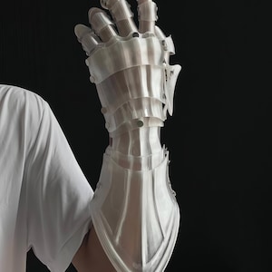 May include: A clear, articulated gauntlet, likely 3D-printed, with a segmented design. The armor-like glove features a series of plates and rivets, with a pointed lower edge. The background is black, and a white shirt is visible in the frame.