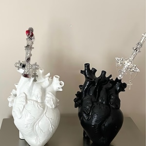 Puede incluir: Dos jarrones con forma de corazón anatómico, uno blanco y otro negro, cada uno adornado con una espada decorativa plateada. El jarrón blanco tiene un acento rojo en la espada. Los jarrones se exhiben sobre una superficie metálica.