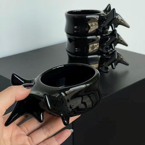 Peut inclure: Tasse en céramique noire brillante avec un design en forme de vertèbre. La tasse a une forme cylindrique avec des éléments pointus et saillants. Une tasse est tenue, tandis que deux autres sont empilées en arrière-plan. Les tasses sont sur une surface noire.