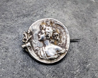 May include: Silver brooch with a detailed design of a woman's profile with a floral accent.