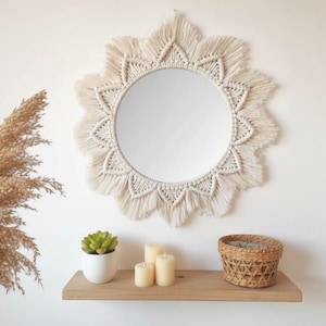 Puede incluir: Espejo redondo de macramé color crema, con diseño de sol. Cuelga de una pared blanca sobre una estantería de madera. En la estantería hay una pequeña suculenta en una maceta blanca, tres velas y una cesta tejida.
