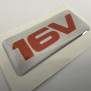 May include: A rectangular silver sticker with the red text "16V" is displayed on a white background. The sticker has a glossy finish and is likely an automotive emblem or decal.