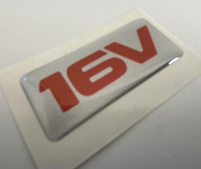 May include: A rectangular silver sticker with the red text "16V" is displayed on a white background. The sticker has a glossy finish and is likely an automotive emblem or decal.