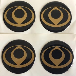 Puede incluir: Cuatro pegatinas circulares dispuestas en una cuadrícula de dos por dos. Cada pegatina presenta un fondo negro brillante con un gráfico dorado centrado que consiste en un círculo sólido rodeado por dos formas curvas y puntiagudas.