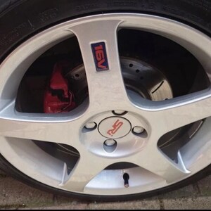 May include: A close-up shot of a silver alloy wheel with a five-spoke design. The wheel has a red brake caliper and a tyre with the word "NEXEN" visible. The centre cap has the letters "RS" in red.