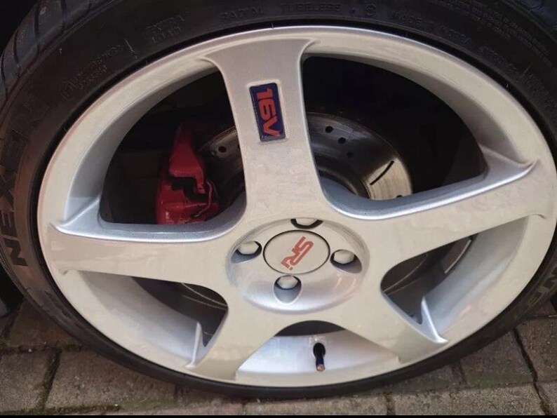 May include: A close-up shot of a silver alloy wheel with a five-spoke design. The wheel has a red brake caliper and a tyre with the word "NEXEN" visible. The centre cap has the letters "RS" in red.