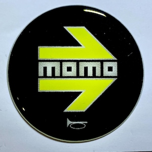 Pode incluir: Um autocolante redondo preto com um design de seta amarelo brilhante. A palavra "MOMO" está em letras prateadas dentro da seta. Um pequeno símbolo de buzina está na parte inferior. O autocolante tem um acabamento brilhante.