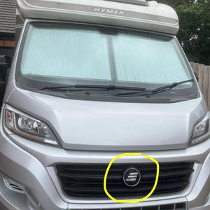 Peut inclure: Vue avant d'un camping-car Hymer argenté avec un pare-brise recouvert d'un store plissé blanc. Une calandre noire est située sous le capot, comportant un emblème circulaire avec trois barres blanches horizontales dans un cercle jaune.
