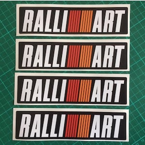 Pode incluir: Quatro autocolantes retangulares com o texto "RALLI ART" em branco sobre fundo preto. Uma faixa vertical de linhas vermelhas, laranja e amarelas separa as palavras. Para entusiastas de carros.
