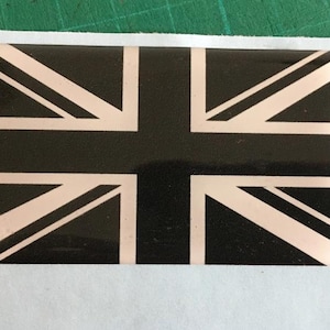 Puede incluir: Una pegatina rectangular con el diseño de la bandera de la Union Jack. La bandera tiene un fondo negro con detalles blancos y rosa claro. La pegatina está sobre un respaldo blanco, con una superficie verde en el fondo.