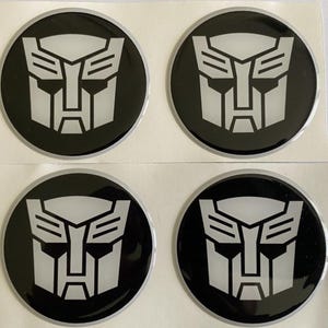 May include: Four black and silver circular decals featuring the Autobot symbol. The Autobot symbol is a stylized robot face in silver against a black background. The decals are on a white backing.