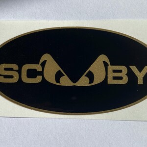 May include: An oval sticker with a black background and gold lettering. The word "SC" is on the left, and "BY" is on the right. A gold-coloured stylized design is in the centre. The sticker is on a white background.