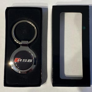 以下が含まれることがあります： 黒い背景に赤と白のRS5ロゴが入った、シルバーのメタルRS5キーホルダー。キーホルダーは、透明な窓が付いた黒い箱に入っています。