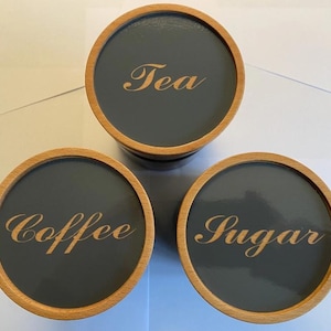 Puede incluir: Tres botes redondos con bordes de madera y tapas grises. Las tapas están etiquetadas con "Tea", "Coffee" y "Sugar" en una fuente decorativa. Los botes están diseñados para el almacenamiento en la cocina.