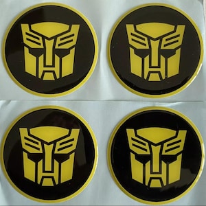 May include: Four circular black and yellow stickers featuring the Autobot symbol. The Autobot symbol is a yellow face design on a black background. The stickers are on a white surface.