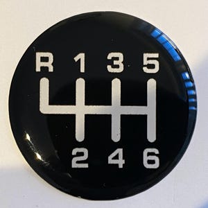 May include: A circular black emblem featuring a white manual transmission gear shift pattern. The design displays the letters R, 1, 3, 5 across the top and 2, 4, 6 across the bottom, connected by vertical and horizontal lines indicating gear positions.