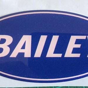 Könnte beinhalten: Ovaler Aufkleber mit dem Namen "BAILEY" in hellrosa Buchstaben. Der Aufkleber hat einen dunkelblauen Hintergrund und einen hellrosa Rand. Der Aufkleber befindet sich auf einer weißen Oberfläche.