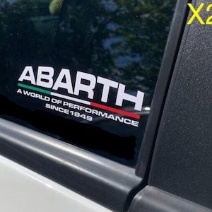 Puede incluir: Calcomanía blanca de Abarth con el texto "ABARTH A WORLD OF PERFORMANCE SINCE 1949". La calcomanía tiene rayas verdes y rojas. La calcomanía está en la ventana de un coche.