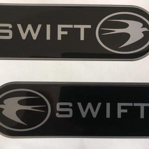 Puede incluir: Dos emblemas rectangulares negros con la palabra "SWIFT" en letras plateadas. Uno tiene un gráfico de pájaro plateado dentro de un círculo. El otro tiene el gráfico del pájaro en el otro lado.