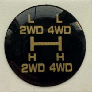 Pode incluir: Um autocolante preto e circular com letras douradas. O texto inclui "L", "2WD", "4WD" e "H", juntamente com uma linha horizontal. O autocolante é provavelmente para o sistema de tração às quatro rodas de um veículo.