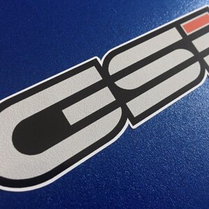 May include: A sticker featuring a graphic design in grey, black, and red. The design includes the letters "GE" in a bold font, with a red rectangular accent. The sticker has a white border and is on a blue background.