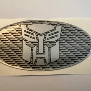 May include: Silver oval emblem featuring the Transformers Autobot symbol. The emblem has a textured background and a glossy finish. The Autobot symbol is a stylized robot face.