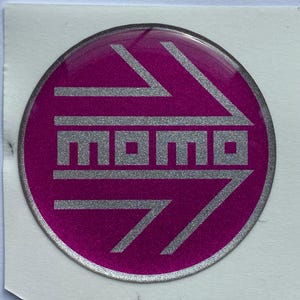 Może przedstawiać: Okrągła naklejka z żywym magentowym tłem i metalicznym srebrnym wykończeniem. Projekt zawiera słowo MOMO pogrubioną, blokową czcionką, otoczone dwiema dużymi, stylizowanymi strzałkami skierowanymi w prawo, wykonanymi w kolorze srebrnym.