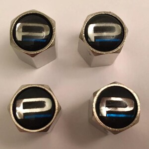 May include: Four silver-coloured tyre valve stem caps. Each cap has a black circular face with a white "P" and a blue stripe. The caps are hexagonal in shape.