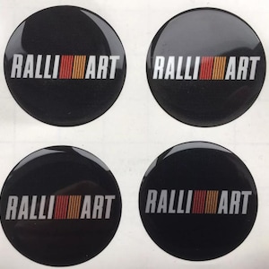 Puede incluir: Cuatro pegatinas negras, brillantes y circulares. Cada pegatina presenta la palabra "RALLI ART" en blanco, en negrita, con fuente sans-serif. Una franja vertical de líneas rojas, naranjas y amarillas está a la derecha de la palabra "RALLI".
