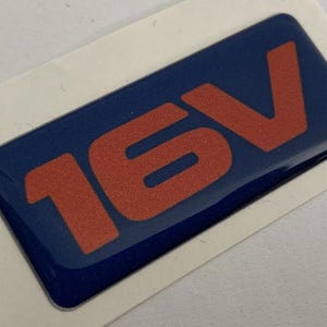May include: A rectangular blue sticker with the text "16V" in orange. The sticker has a glossy finish and is on a white background.