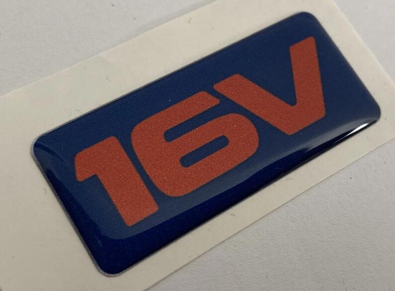 May include: A rectangular blue sticker with the text "16V" in orange. The sticker has a glossy finish and is on a white background.