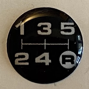 Pode incluir: Um botão de mudança de marcha preto com um padrão prateado indicando as posições das marchas: 1, 3, 5, 2, 4 e R (marcha a ré). Os números e a letra estão em uma fonte sem serifa. O botão é redondo e tem um acabamento brilhante.