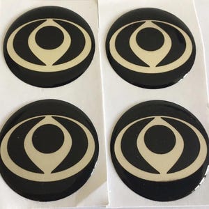 Puede incluir: Cuatro pegatinas circulares negras y crema. Cada pegatina tiene un fondo negro con un diseño crema en el centro. El diseño es una forma de ojo estilizada con un círculo central y líneas curvas.