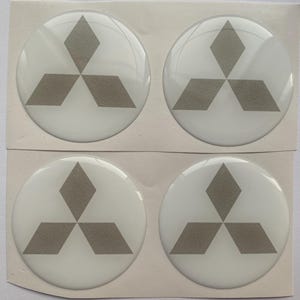 May include: Four white, circular car emblem stickers with a gray Mitsubishi logo. The logo features three gray diamond shapes arranged in a triangular pattern, with one diamond at the top and two at the bottom.