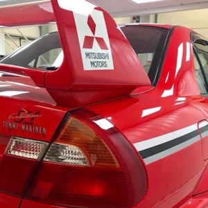 Puede incluir: Un coche rojo Mitsubishi Lancer Evolution Tommi Mäkinen Edition. El coche presenta un gran alerón trasero con el logotipo de Mitsubishi Motors en blanco. El coche tiene una carrocería roja con rayas negras y plateadas.