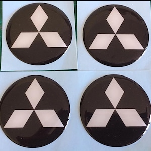 May include: Four circular black emblems with a white three-diamond logo. The emblems are glossy and appear to be stickers or decals. The background is a light green surface.