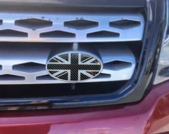 Peut inclure: Calandre de voiture avec un emblème Union Jack. L'emblème ovale présente un motif en fibre de carbone et le drapeau britannique. La calandre est argentée, et l'avant de la voiture est rouge.