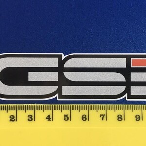 May include: A rectangular sticker featuring the letters "GSI" in a stylized font. The letters are mainly grey with black accents and a red rectangle. A yellow ruler with mm and cm markings is at the bottom.