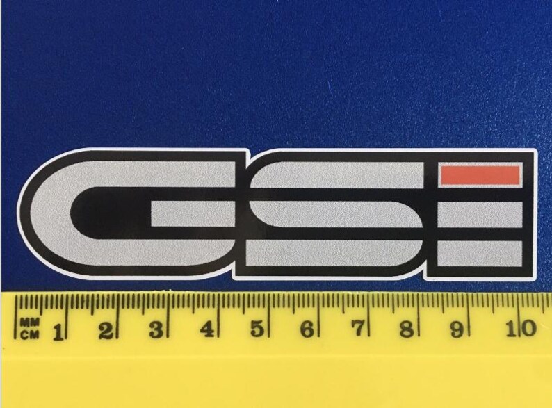 May include: A rectangular sticker featuring the letters "GSI" in a stylized font. The letters are mainly grey with black accents and a red rectangle. A yellow ruler with mm and cm markings is at the bottom.