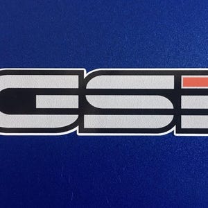 May include: A sticker featuring the letters "GSI" in a stylized design. The letters are silver with black and white outlines, and a small orange rectangle. The sticker is on a blue background.