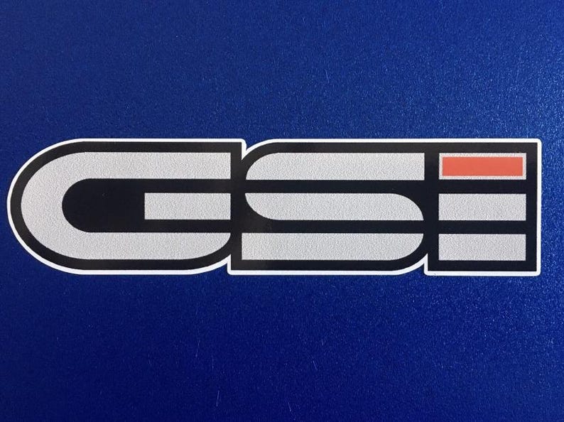 May include: A sticker featuring the letters "GSI" in a stylized design. The letters are silver with black and white outlines, and a small orange rectangle. The sticker is on a blue background.