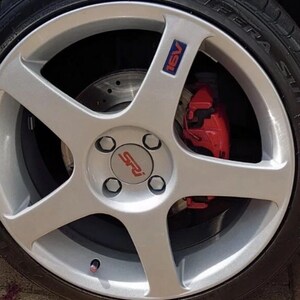 May include: A silver alloy wheel with a five-spoke design. The wheel features a red brake caliper and a tire. The center cap has the letters "SRI" in red. The wheel also has a blue and red "16V" emblem.